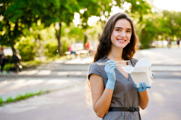 医学手袋とマスクに巻き毛のある若い驚きの女の子は手と笑顔で箱の中にwokを保持しています.白箱配送のうどん。検疫covid 19での日本食レストランの広告 - 写真・画像