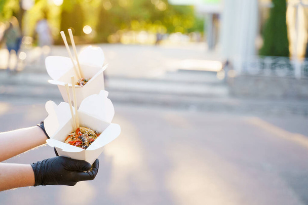 Detailní chutné jídlo wok v krabici dodávky. Muž drží udonové nudle s tempuru, krevetami, v rukou v černých lékařských rukavicích. Čínské kořeněné jídlo v jednorázových ekologických krabicích. 2 Wok box udon inzerovat 1 + 1 - Fotografie, Obrázek