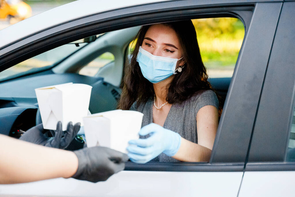 Young attractive girl in car gets 2 wok in box udon noodles with tempuru, shrimps, in hands and smiles. Courier in medical gloves. Udon noodles in white box delivery. Wok box udon advertise 1+1. - Photo, Image