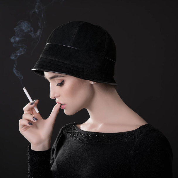 Young fashion woman smoking cigarette in hat and black drees over dark background. Vintage female portrait, styling. Image toned. - 写真・画像