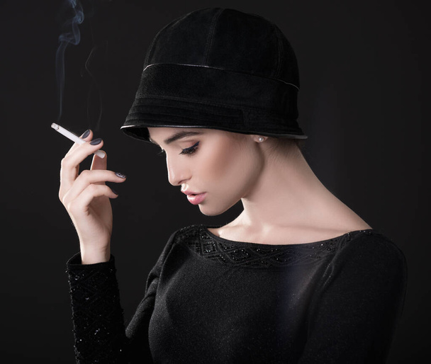 Young fashion woman smoking cigarette in hat and black drees over dark background. Vintage female portrait, styling. Image toned. - Photo, Image