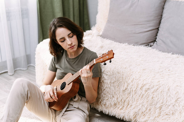Piękna kobieta gra na gitarze w domu. Młoda dziewczyna gra w ukulele podczas samoizolacji. - Zdjęcie, obraz