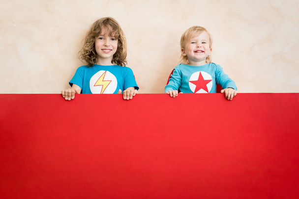 Dzieci superbohaterów trzymające czerwony sztandar puste. Zabawne dzieciaki bawią się w domu. Koncepcja snu i wyobraźni - Zdjęcie, obraz