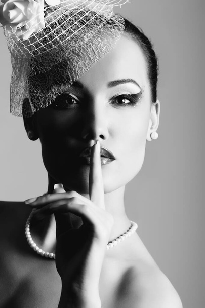 Pretty lady in white hat with veil and roses, retro glamour beauty portrait. Beautiful woman with perfect makeup looking at camera with shhh gesture, high fashion look. Girl with a pearl earring, black and white - Foto, afbeelding