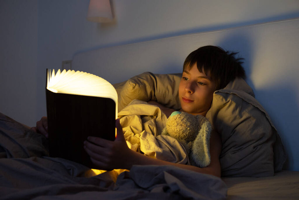 Adorable boy reads book before sleeping in bed at home with his old toy. Happy childhood. - Zdjęcie, obraz