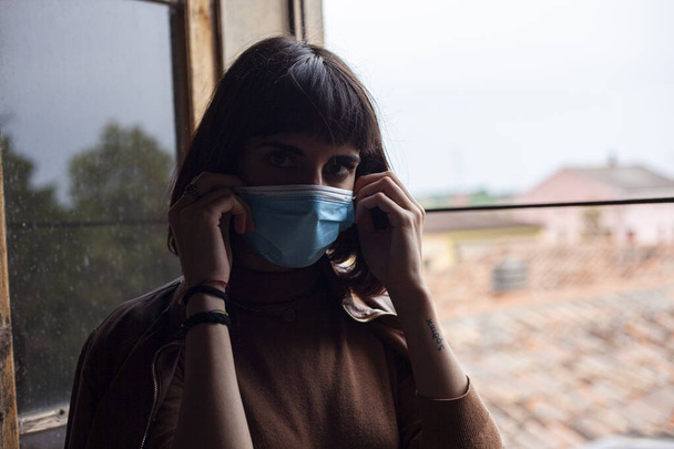 Meisje met medisch masker bij het raam in haar huis tijdens covid quarantaine periode - Foto, afbeelding
