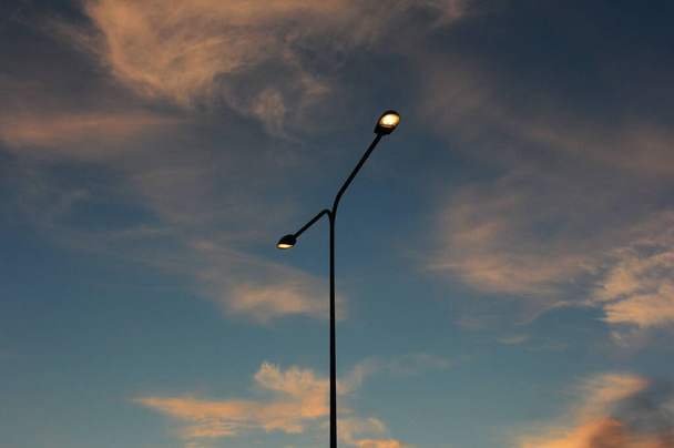 Улица освещения фонарный столб с двумя огнями против облачного неба освещается заходящим солнцем
 - Фото, изображение