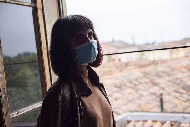 Girl with medical mask at window in her home during covid quarantine period - Fotó, kép
