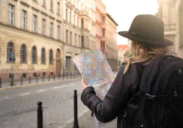 young girl is looking at a city map on the street on a journey.woman has blond hair wearing a leather jacket, hat and backpack.new travel concept in new realities. - Zdjęcie, obraz
