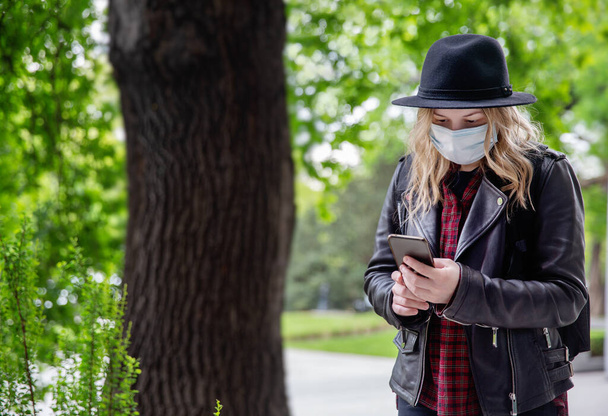 A young woman with blonde hair in a black hat and a medical mask looks at the phone on the street. New realities and safety requirements in a pandemic. the need for a mask while in  society. - Фото, изображение