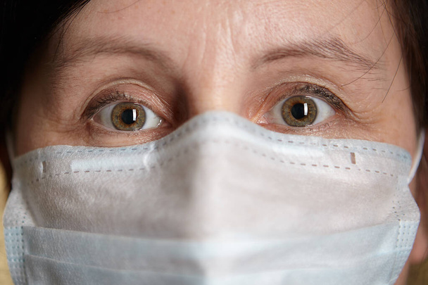 Woman with medical facial surgical mask on her face, her eyes express anxiety and fear because of home isolation during coronavirus infection covid-19 pandemic. - Photo, Image