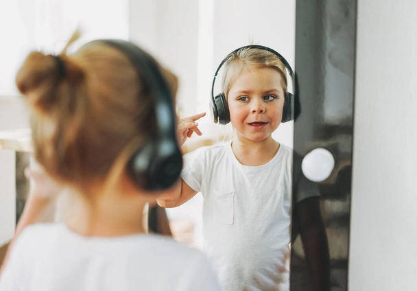 Χαριτωμένο κοριτσάκι με ξανθά μαλλιά σε ακουστικά κοιτάζοντας στον καθρέφτη - Φωτογραφία, εικόνα