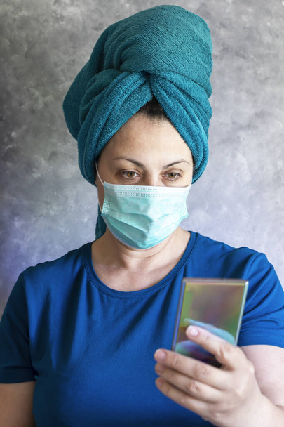 A woman in a medical mask works remotely from home in the kitchen. Conversation on mobile communications. Distance learning online, training and work at home. On a gray background. - Photo, Image