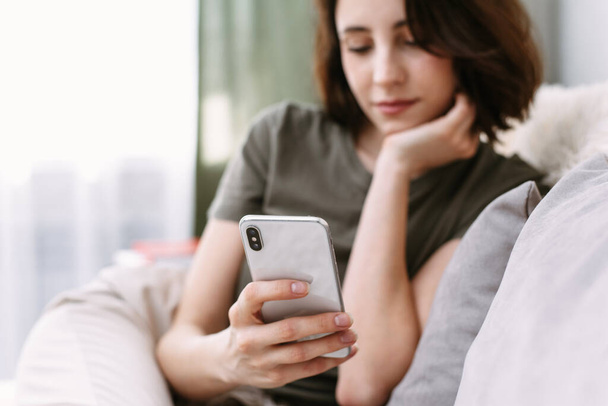 Femme attrayante utilisant un smartphone tout en étant assis sur le canapé à la maison. Shopping et réseautage social par téléphone
. - Photo, image