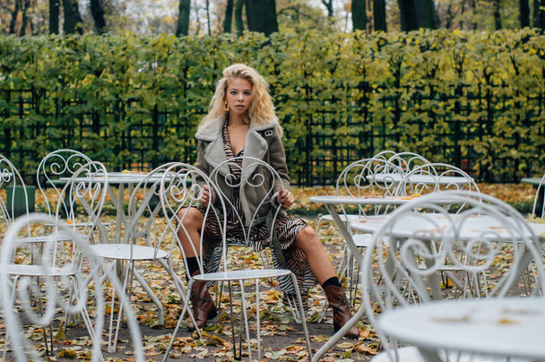 Nuori blondi tyttö istuu pöydässä julkisessa puistossa syksyllä ja katselee kameraan
 - Valokuva, kuva