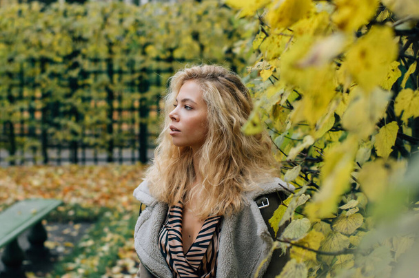 Chica rubia joven que se queda cerca de los árboles en el parque público en otoño y mirando a un lado
 - Foto, Imagen