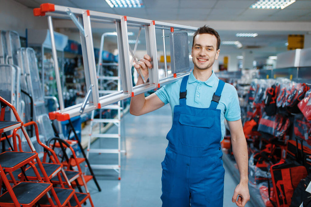 Muž v uniformě drží nové hliníkové žebříky v obchodě s nářadím. Oddělení se žebříky, výběr vybavení v železářství, nástrojárna - Fotografie, Obrázek