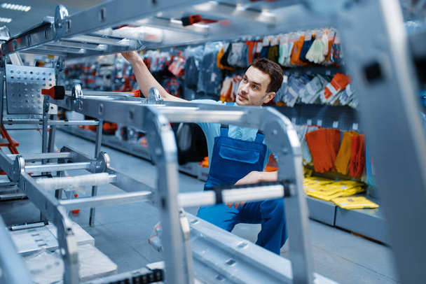 Mannelijke werknemer in uniform houdt nieuwe aluminium trapladders in gereedschapswinkel. Afdeling met ladders, keuze aan apparatuur in de ijzerwarenwinkel, instrumentensupermarkt - Foto, afbeelding