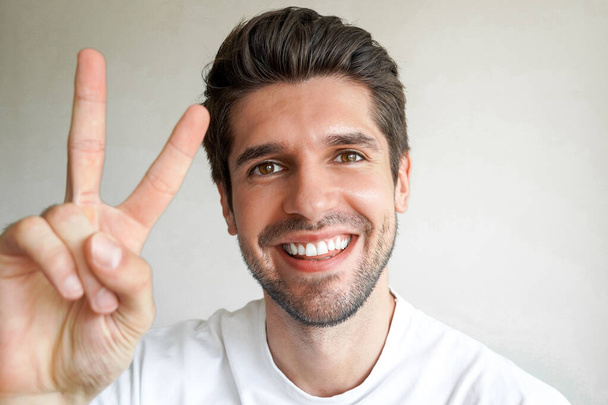 Portrait of young handsome funny guy who is showing victory sign by his arms and joyful emotions on his face - Photo, Image