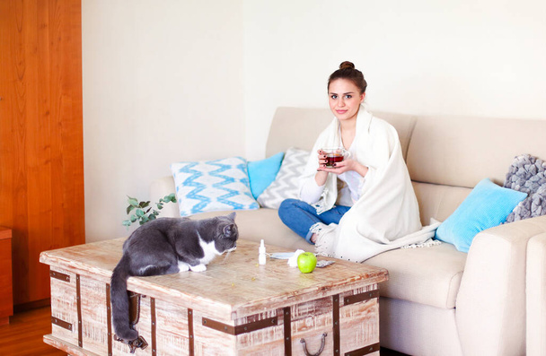 Ill female with cup of hot tea touching forehead with closed eyes while sitting cross legged on couch near table with cat at home - Foto, Imagem