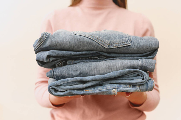 Young shopkeeper woman holding pile of jeans over light background in her hands. Close up - Φωτογραφία, εικόνα