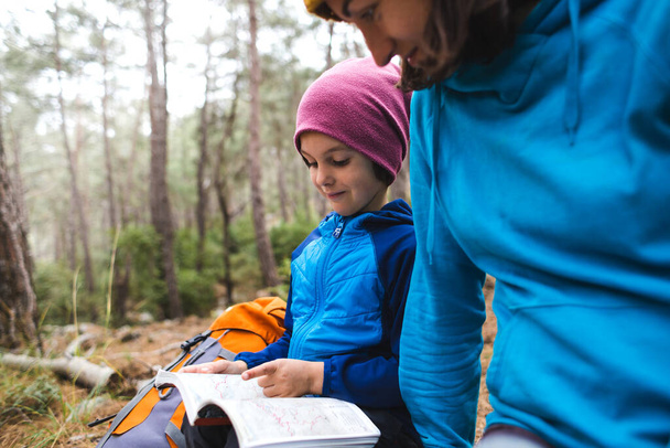 Мальчик с матерью в походе, ребенок смотрит на карту, путешествуя с детьми, ребенок учится определять местоположение на карте, мальчик с рюкзаком походы в лес
. - Фото, изображение