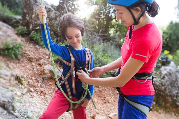 Инструктор учит ребенка пользоваться альпинистским снаряжением. Мальчик готовится забраться на скалу. Женщина показывает ребенку, как использовать карабин для страховки. Мать привязала веревку к системе безопасности. - Фото, изображение