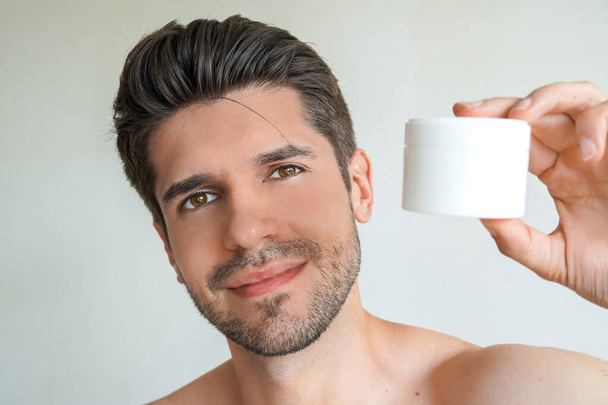 Νέος όμορφος άντρας με τέλειο δέρμα και μαλλιά δείχνει ένα προϊόν στο μπάνιο του. - Φωτογραφία, εικόνα