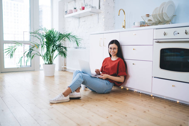 Pleased young stylish woman in red t shirt and denim sitting on floor leaning on white kitchen counter surfing laptop at home looking at camera  - Photo, image