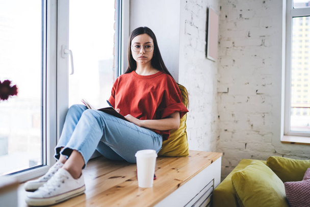 Mindful woman wearing casual clothes writing notes in notebook while sitting on windowsill next to flowers in modern apartment looking away - 写真・画像