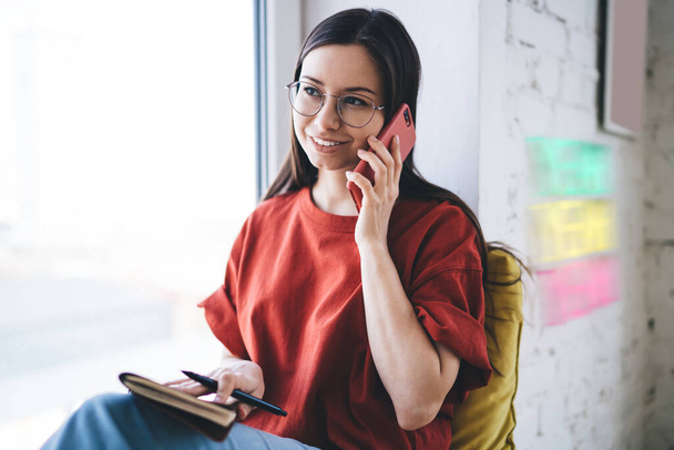 Cheerful woman in casual clothing and glasses having conversation on mobile phone while holding notebook and sitting on window sill - Photo, Image