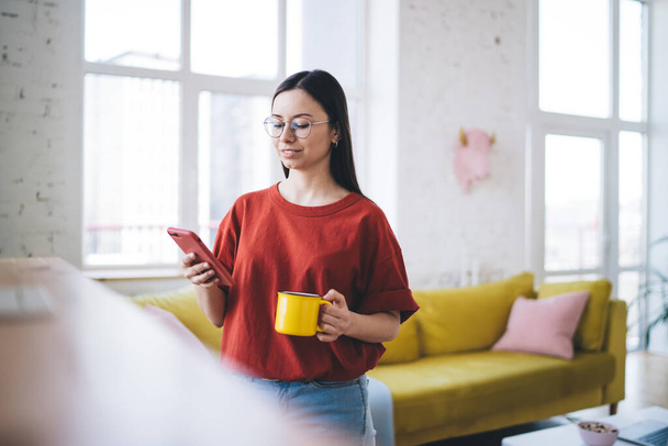 Female in casual clothing and glasses having morning cup of coffee and checking messages on mobile phone on blurred background - Photo, image