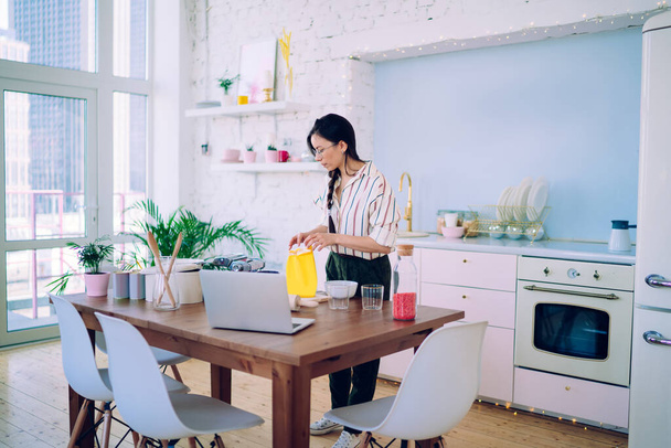 Tranquil lady standing in front of table in kitchen and preparing products and equipment for cooking while watching video with recipe  - Foto, imagen