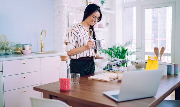 Joyful female in casual clothing having fun while preparing food on wooden table with laptop in kitchen of modern apartment - Foto, afbeelding