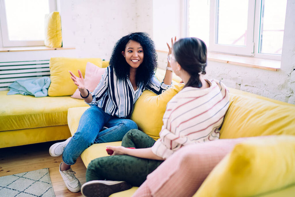 African American woman with delightful smile gesturing while telling exciting story to female friend relaxing on yellow cozy sofa at home - Фото, изображение