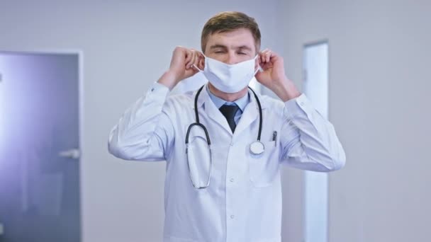 Doctor man wearing the a protective mask in the modern hospital corridor and looking straight to the camera he have a serious face - Кадри, відео