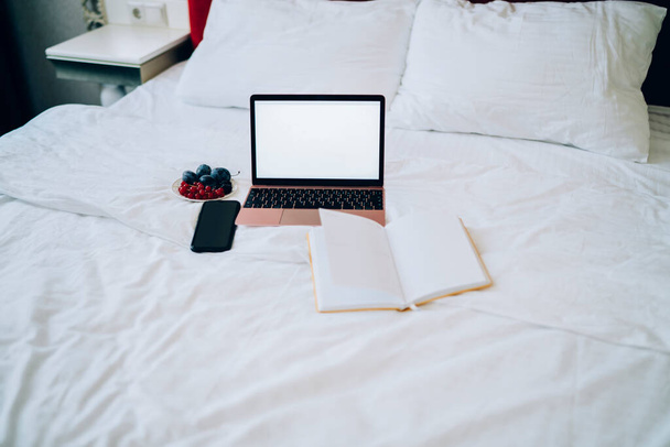 From above open laptop book smartphone and bowl with berries and fruits on bed with pillow and crumpled white linen - 写真・画像