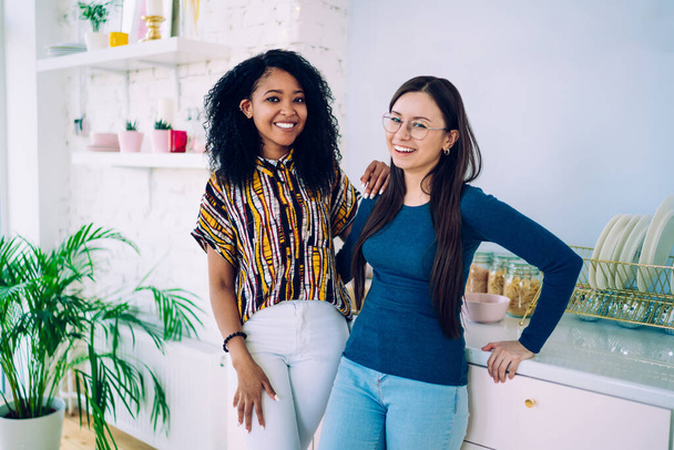Happy young attractive multiethnic women in trendy clothes laughing joyfully and smiling while standing close together in kitchen leaning on cupboard and looking at camera - Foto, Bild