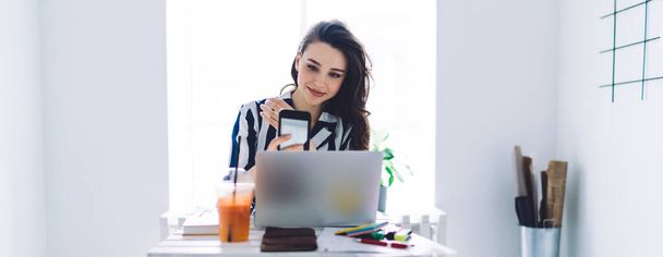 Attractive long haired female worker in striped white blue clothes using smartphone sitting at comfortable workplace in small office room - Фото, зображення