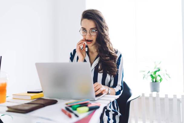 Young attractive woman in glasses and striped dress sitting amazed at desk and touching face with hand while using and looking at laptop - Photo, image