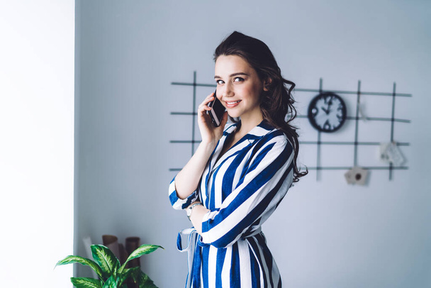 Charming successful swirl haired female entrepreneur in loose striped dress standing at window and arranging for meeting with colleague on mobile phone smiling and looking at camera   - Foto, Imagen