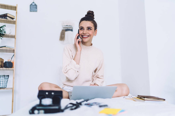 Smiling adult lady in casual clothes making phone call and looking away while sitting crossed legs at laptop on bed against white wall with stationery in light bedroom - Foto, afbeelding