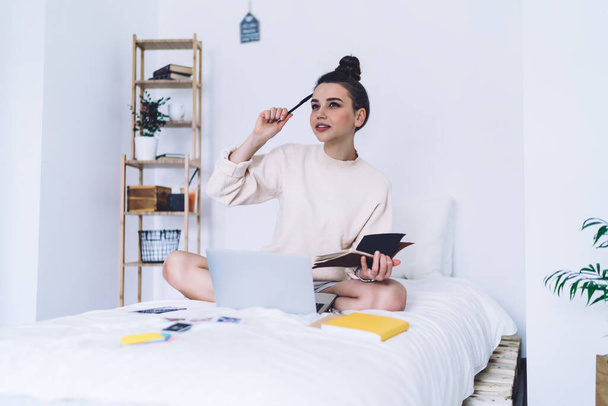 Thoughtful young female startupper sitting with pencil in hand near head and legs crossed on bed elaborating an ideas with books and laptop at loft bedroom  - Photo, image