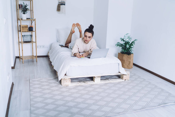 Dark haired modern female with bun in casual clothes lying on bed with laptop and writing notes in cozy small bedroom - Fotoğraf, Görsel
