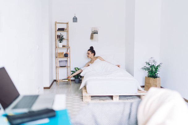 Young female with dark hair wrapped in high bun sitting in bed after waking up and reaching out for phone on shelf - Foto, Imagem