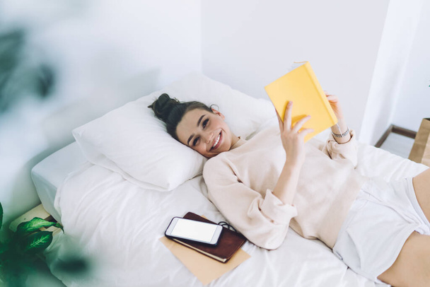 From above of charming young woman lying on single bed in light room with open book in hands smiling widely and looking at camera - Foto, Bild