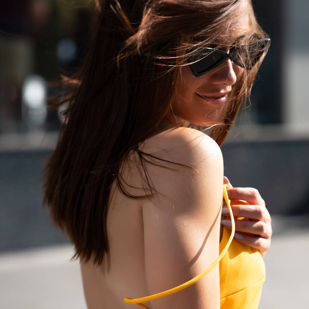 Fashionable pretty stylish girl posing outside. Street fashion. Beautiful young brunette smiling woman wearing yellow dress and black sun glasses walking in city center, summer outdoor.  - Foto, imagen