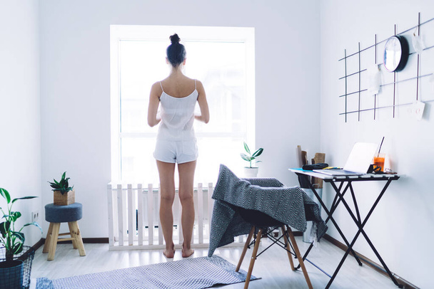 From behind barefoot calm woman with hairbun in pyjamas standing and looking out window in modern apartment with white walls - Foto, afbeelding