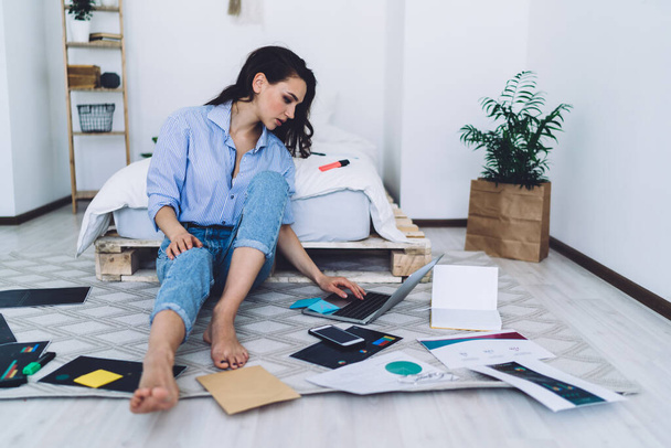 Concentrated serious woman in casual outfit taking notes and browsing laptop while sitting barefoot on floor with graphs in modern room - 写真・画像