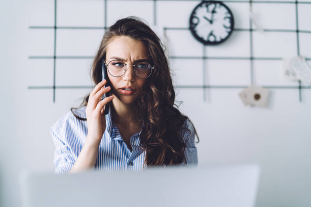 Stressed young female worker in glasses in striped blue shirt listening on mobile phone attentively squinting aside at workplace in office - Foto, immagini